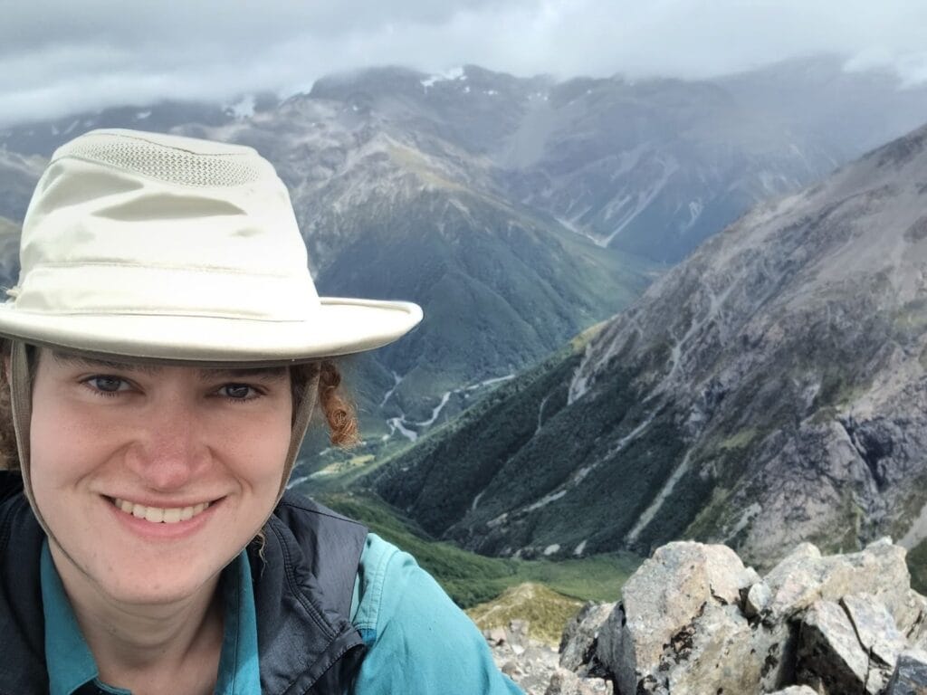 Gwen is pictured on a mountain-hike, she is smiling at the camera and there are more mountains [Southern Alps] behind her.