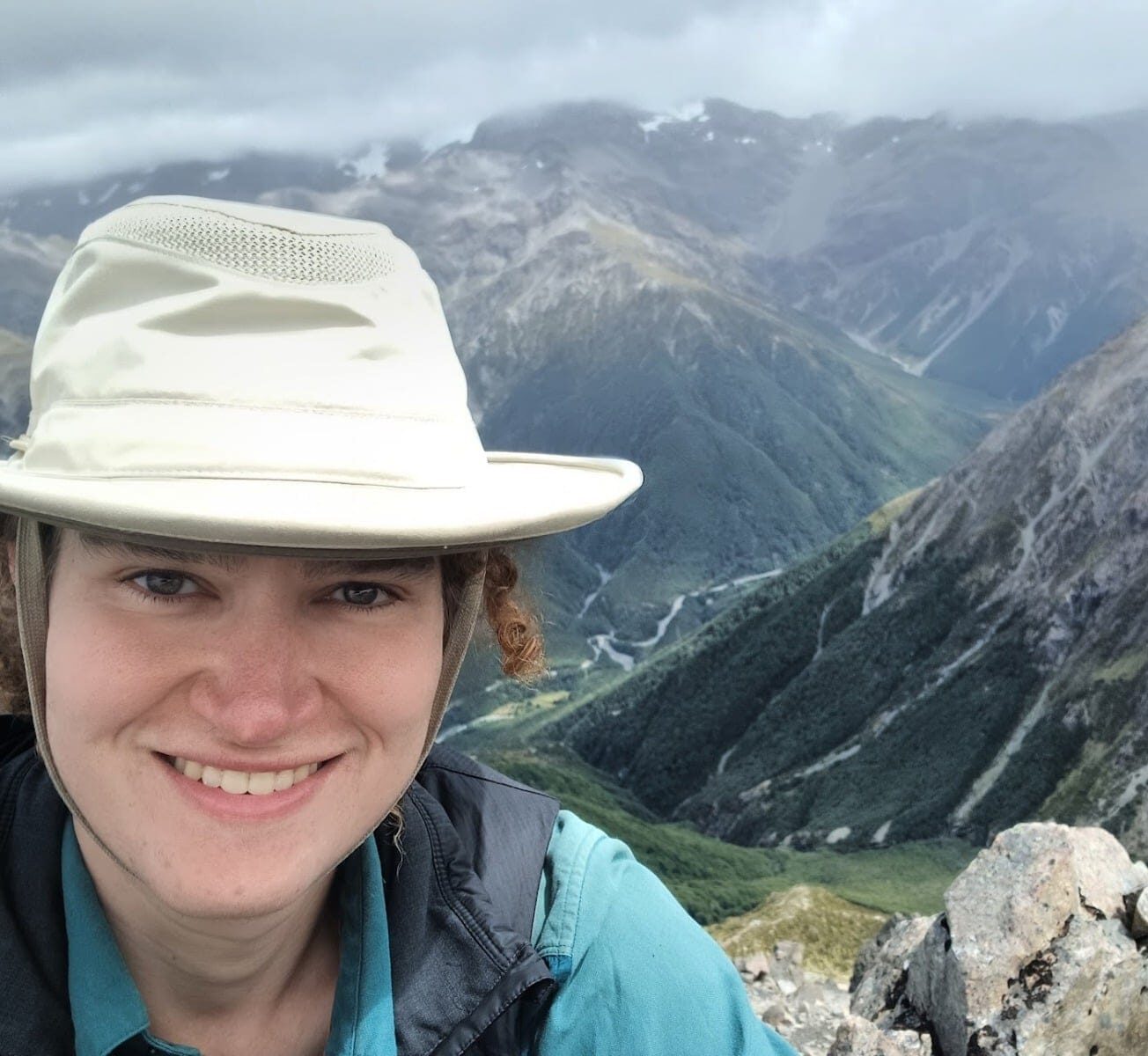 Gwen is pictured on a mountain-hike, she is smiling at the camera and there are more mountains [Southern Alps] behind her.